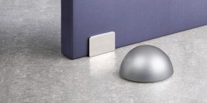 Tope de puerta Inofix 2034 con retenedor magnético en acabado cromo mate, instalado sobre suelo gris con puerta azul, ideal para mantener puertas abiertas sin golpes